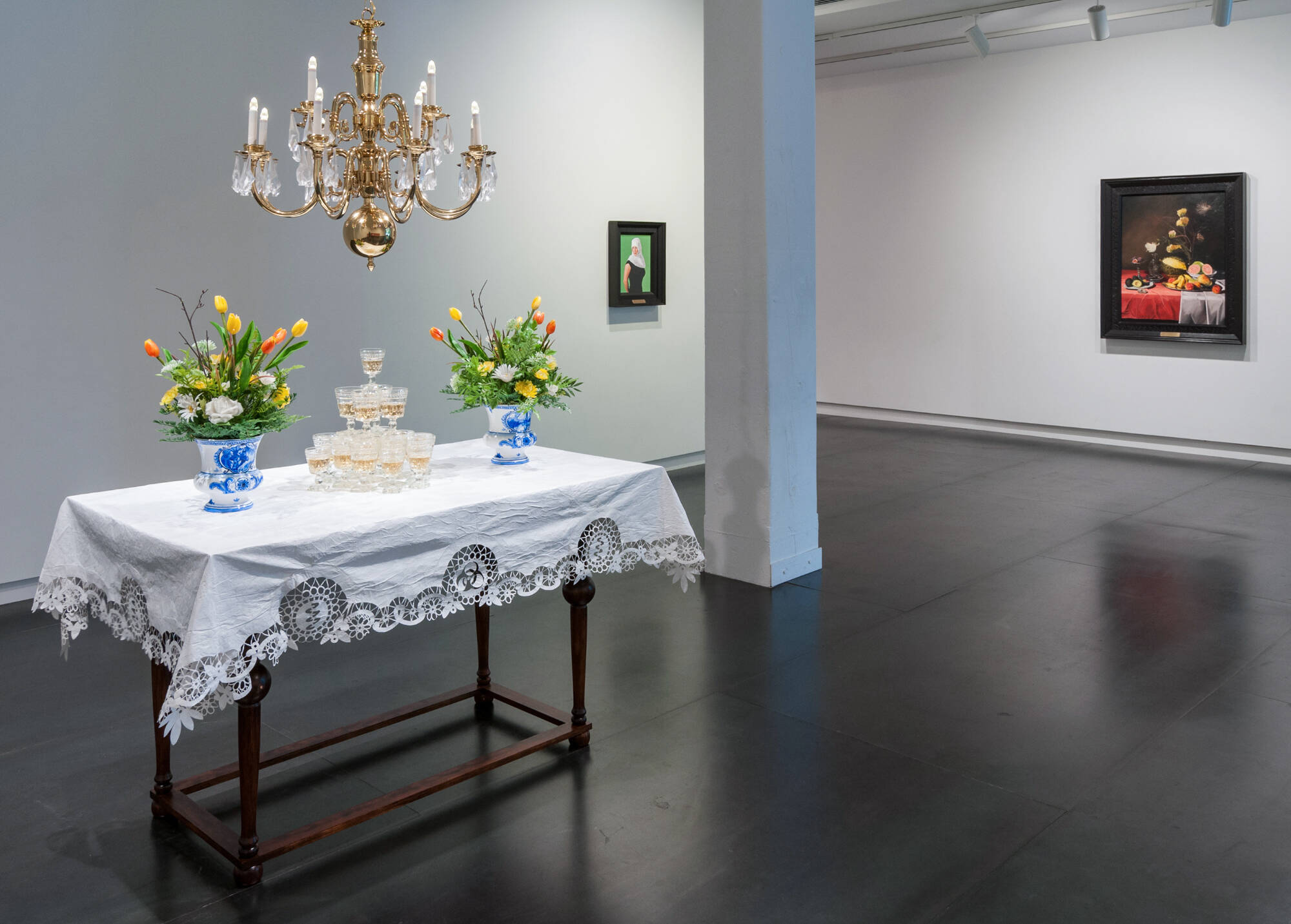 An art installation of a table covered by a white tablecloth, with stacked champaign glasses and two floral arrangements on it. A chandelier hangs over the table.